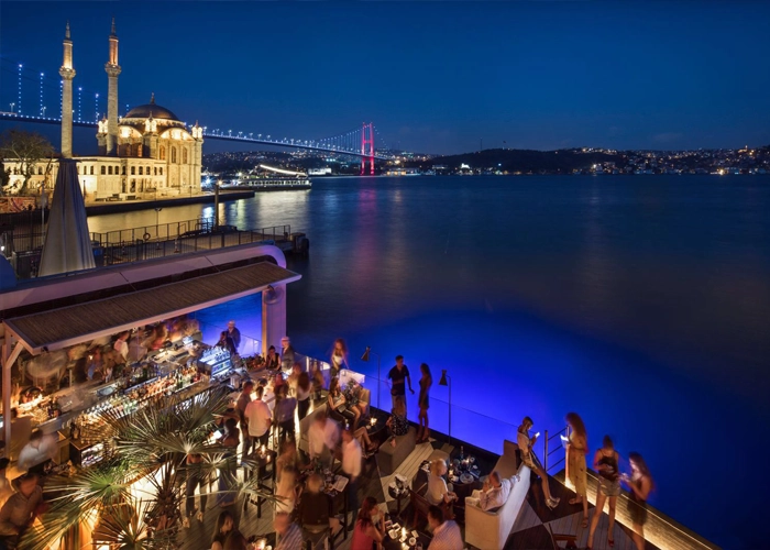 اورتاکوی (Ortaköy) – ویو خفن و حال‌وهوای خاص
اورتاکوی یکی از خاص‌ترین لوکیشن‌های استانبوله، بیشتر به خاطر ویوی فوق‌العاده‌اش به پل بسفر. اینجا بیشتر برای قدم زدن، عکس گرفتن، نشستن کنار آب و لذت بردن از فضا معروفه.
عصرها و شب‌ها بهترین زمان رفتنه، وقتی نور پل روشن میشه، کل فضا یه حال‌وهوای متفاوت می‌گیره. همین باعث شده یکی از محبوب‌ترین جاها برای تجربه شب‌های استانبول باشه.
اینجا بیشتر برای تفریح کوتاه و حال کردنه، نه زندگی روزمره. قیمت‌ها نسبت به چیزی که می‌گیری یه مقدار بالاست، ولی ویو و فضا تا حد زیادی جبرانش می‌کنه.
